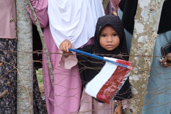 A young girl waits to see Indo<em></em>nesian President Joko Widodo. She is wearing a black headscarf and waving an Indo<em></em>nesian flag