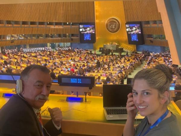 Lisa is sitting in the UN General Assembly with Grant Lindsay, the President and Chair of the Australian Federation of Disability Organisations. Grant is wearing a black jacket and has an ear piece on his ear. Lisa is wearing a green dress and is holding an ear piece to her ear.