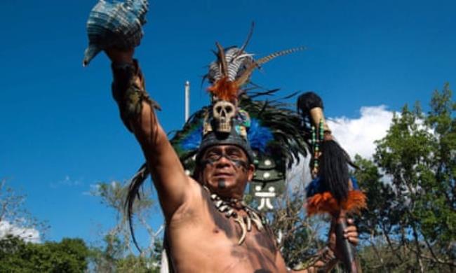 A man in traditio<em></em>nal Maya costume, with a feathered skull adorning his head, holds aloft a vessel in one hand, with a black staff in the other