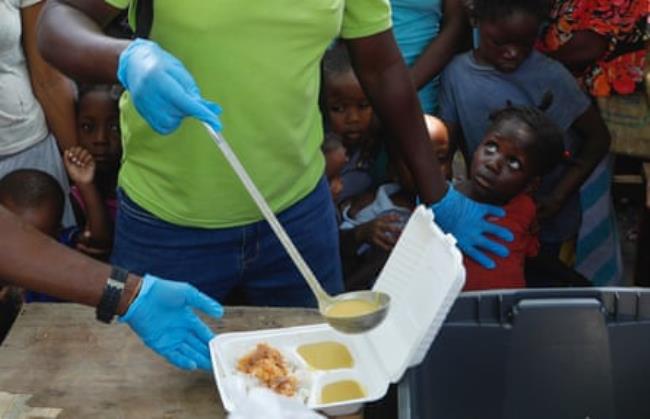 Someone ladles food into a plastic container, while children look on behind them
