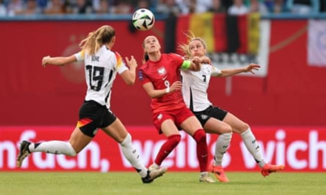 Ewa Pajor, in a red kit playing for Poland, goes to head the ball with Klara Buehl and Kathrin Hendrich of Germany, in white shirts and black shorts, challenging her from either side. She has long light brown hair which is tied back, while the two German women have long blond hair in ponytails. 