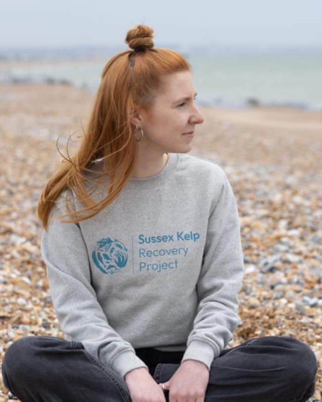 A woman sits crossed-legged on a shingle beach, her shirt has the words Sussex Kelp Recovery
