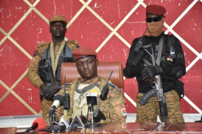 Ibrahim Traoré in military uniform, sitting a desk, with two soldiers standing behind him