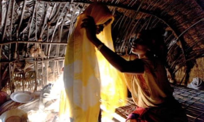 African woman with her daughter in a traditio<em></em>nal hut. The woman is seated, the girl standing, and the sun streams through the door.