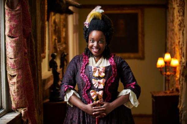 Lolly Adefope as Kitty in Ghosts, wearing an old-fashio<em></em>ned Georgian-era style dress and feather in her hair