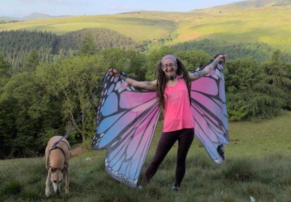 A woman stands on a beautiful hillside wearing butterfly wings and standing beside a dog