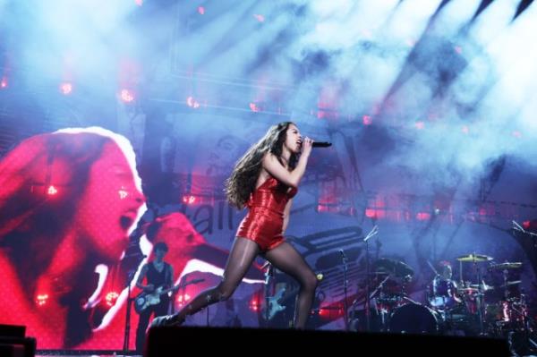 Olivia Rodrigo performs on stage during Guts World Tour. She scrunches up her face as she sings into microphone, wearing red bodysuit.