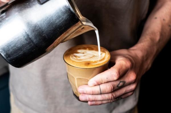 Close up of person with tattooed finger pouring milk from text<em></em>areal jug into glass of cafe latte.