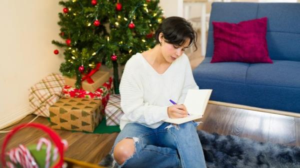 Young woman sitting on the floor in her living room, writing her christmas wish list in a notebook