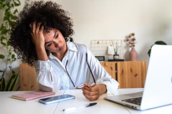 Woman sits at her desk looking stressed while staring at a laptop and holding her glasses in her hands