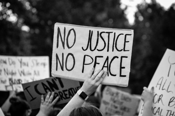 A black-and-white photo shows a protester holding up a No Justice, No Peace sign.