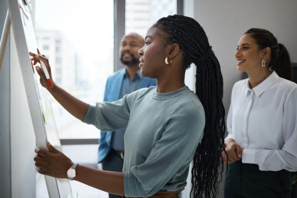 A Black woman writing on a whiteboard while a man and a woman look on