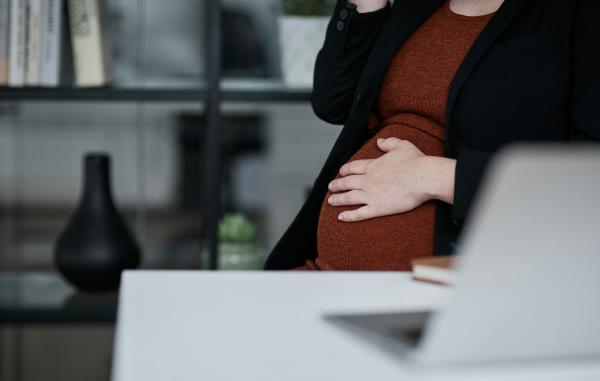 Close up of the pregnant stomach of a person sitting in front of a desk with an open laptop on it