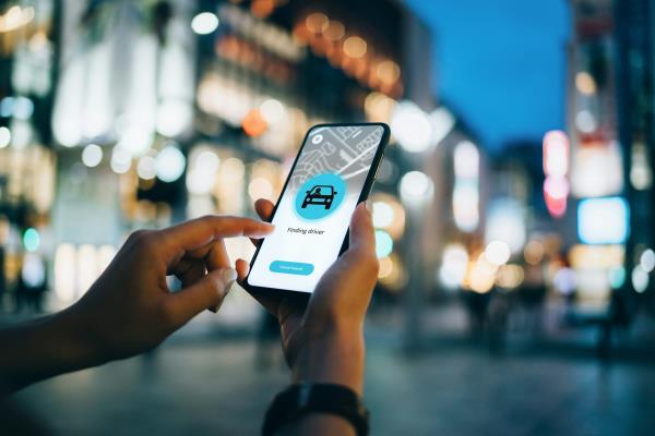 A close-up of a young woman using an app on smartphone to arrange a ride in downtown city street, with an illuminated city traffic scene as background.