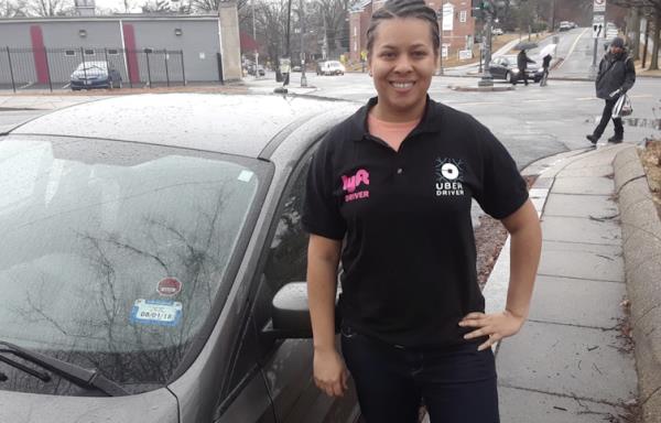 A smiling professor stands in front of a car, her hand on her hip. She is dressed all in black, and her shirt has the Uber and Lyft logos.