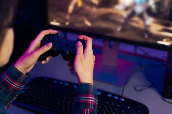 Close up of a pair of hands using a video game co<em></em>ntroller to play a game that is seen on a computer screen in the background