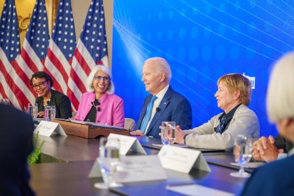 President Joe Biden sits at a table with three others with American flags serving as a background.