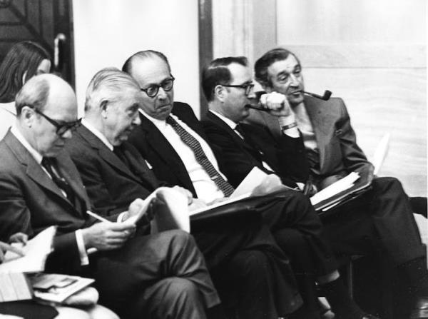 Five seated men in suits holding papers and conversing