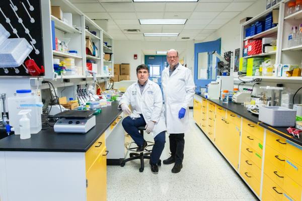 Two men wearing lab coats and gloves standing in a lab with stern looks on their faces. One is seated and they are surrounded by the counters and shelves of a lab