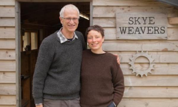 Roger and Andrea Holden stand in front of the doorway of a wooden building with a sign that reads Skye Weavers. Roger is tall with white hair and Andrea has very short dark hair; they are both wearing woollen jumpers. 