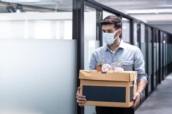 A man in a mask carrying a box of belo<em></em>ngings past a row of cubicles