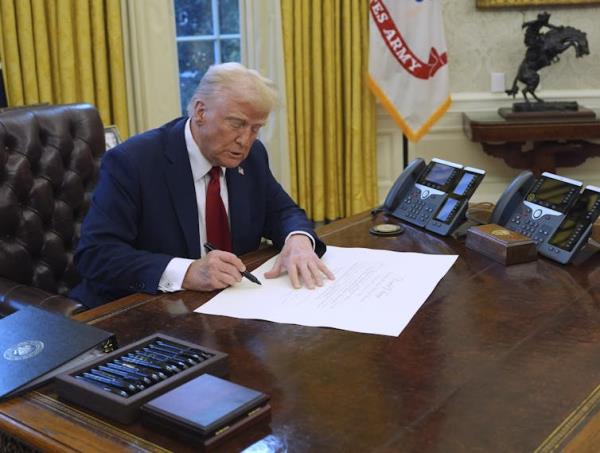 A man with fluffy greyish-blond signs a new<em></em>line at a desk in an ornate office.