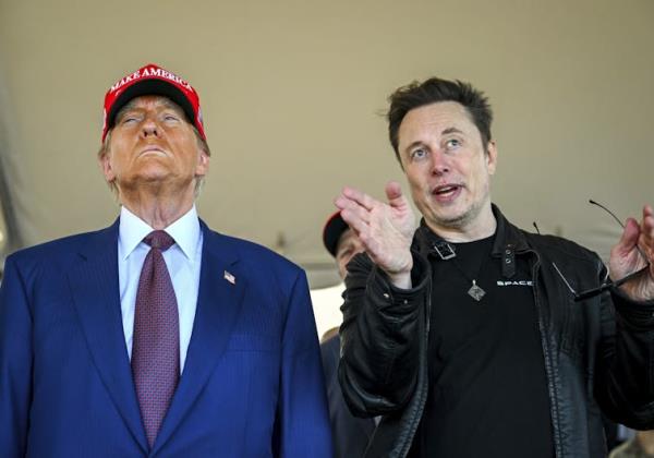 An older man with heavy facial bro<em></em>nzer wearing a red ball cap stands next to a younger man with dark hair who gestures as he looks ahead.