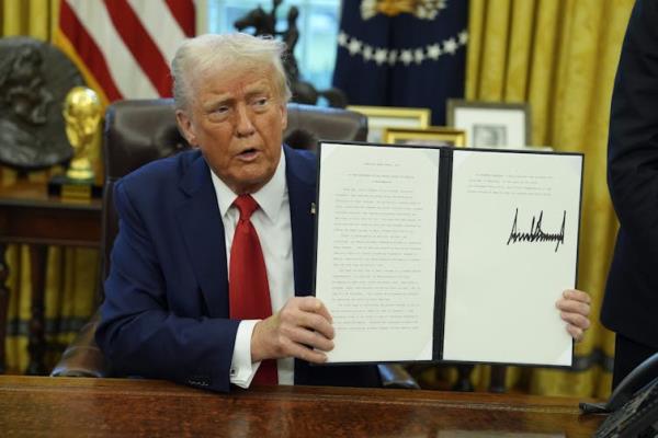 A white man with orange-tinged skin and white hair holds up a signed portfolio