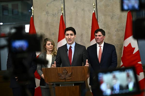 A white man in a suit speaks from a podium with Canadian flags standing behind him. He is flanked by a white woman and a white man