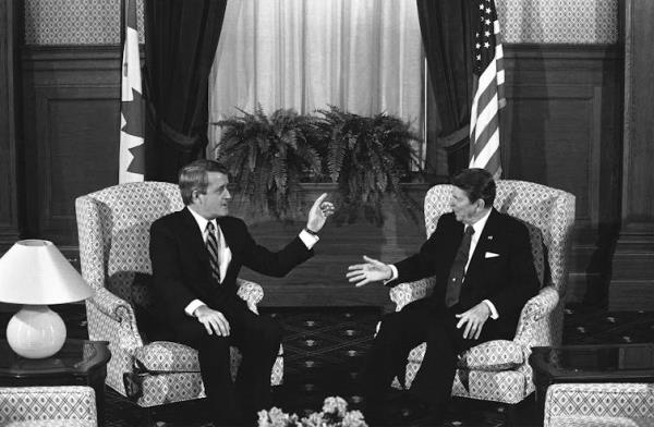 Black and white photo of two white men sitting in armchairs and havin<em></em>g a conversation. A Canadian flag stands behind one and and American flag stands behind the other