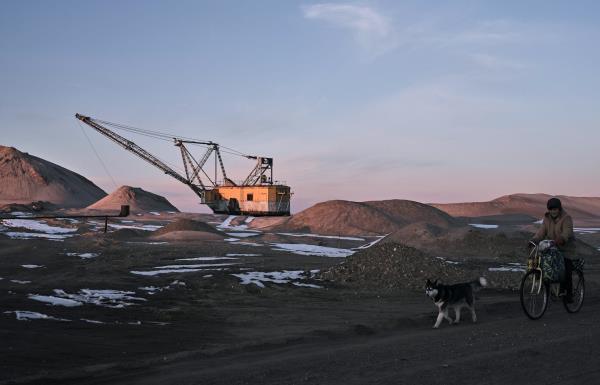 A person on a bicycle with a dog rides past an open mining area with a large excavator in the distance.